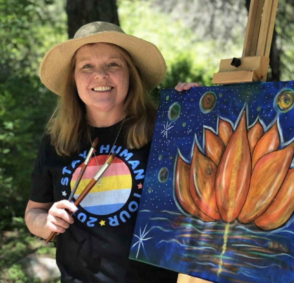 Karen Gillette holding paintbrushes next to a vibrant painting of a lotus flower under a starry sky
